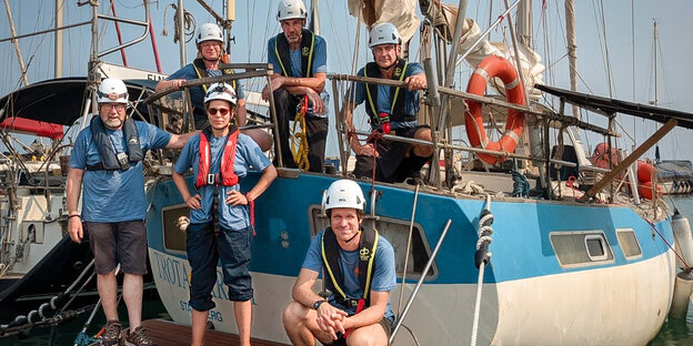 Eine Crew posiert vor einem kleinen hellblau-weißen Segelschiff