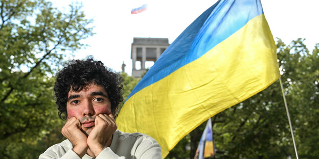 junge r Mann neben ukrainischer Flagge, im Hintergrund die russische Botschaft in Berlin