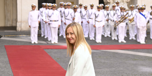 Prime Minister Giorgia Meloni, with blonde hair and a white jacket, in front of a chapel.