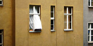 Das Bild zeigt eine Wand eines Mietshauses mit mehreren Fenster und einem wehenden Vorhang.