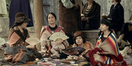 Auf dem Boden sitzt eine Gruppe Frauen, die Kleidung ein Mix aus indigenen Stoffen und Kleidern der 1920er Jahre