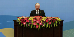 Präsident Putin steht vor einem breiten Pult, das mit Blumen behangen ist.