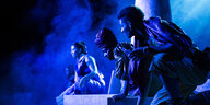 Vier Schwarze Menschen hocken in blauem Licht auf einer Bühne
