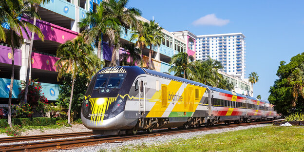 Ein bunter Zug fährt auf Schienen vor der bunten Wand einer Wohngebäudereihe in West Palm Beach in Florida, dahinter steckt ein Wohnhochhaus.
