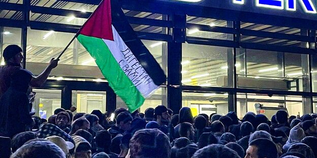 Eine Menschenmenge drängt sich vor dem Eingang des Flughafens in Dagestan, ein Mann schwingt eine große palästinensische Flagge