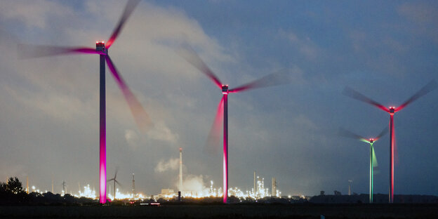 Wind turbines in front of an illuminated refinery.