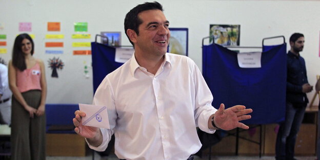 Alexis Tsipras hält lächelnd seinen Stimmzettel in der Hand, im Hintergrund Wahlkabinen