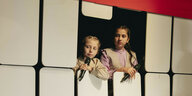 Zwei Mädchen mit Mikrafonen in der Hand schauen aus dem Bühnenbild der Produktion "Land aller Kinder"