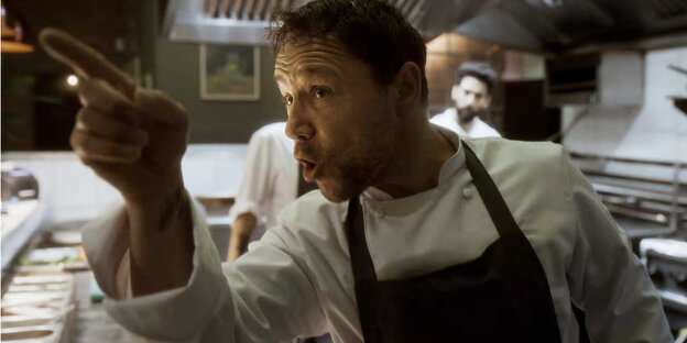 Szene in einer Restaurantküche: Ein Koch mit einer blauen Schürze zeigt mit dem Finger auf jemanden außerhalb des Bildausschnitts. Er schaut drohend zu der anderen Person und schimpft.