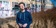 Benjamin Fredrich im blauen Hoody steht vor einem Flachbau, der mit Graffiti besrpüht ist und kneift ein Auge zu