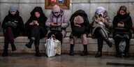 Frauen sitzen in einem U-Bahnhof nebeneinander, alle schauen auf ihre Handys