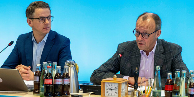 Carsten Linnemann and Friedrich Merz are sitting at a table;  In front of them are many bottles of water and cola and in the background a sky blue wall.
