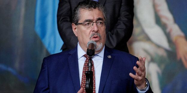 A man in a dark blue suit, Bernardo Avelaro, stands in front of the microphone.