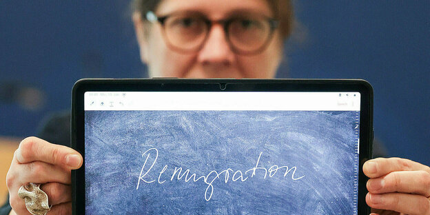 A woman holds a tablet in front of the camera.  On it you can see the words Remigration.