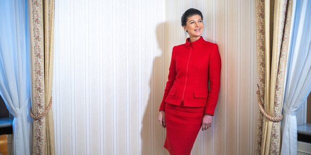 Sahra Wagenknecht wears a red suit and stands in front of the textile wallpaper of a hotel room.