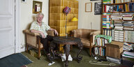 Ein alter Mann sitzt am gelben Kachelofen, viele Bilder und Bücher im Zimmer