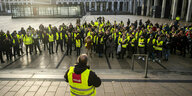 Streikende mit gelben Verdi-Westen stehen vor dem Flughafen BERlin Brandenburg BER, vor ihnen steht ein Mann, auch mit Weste und Mikrofon