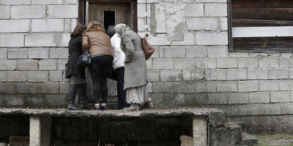 Four women stand on a ramp and look through a half-open door;  the house looks dilapidated