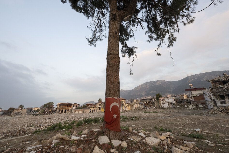 Ein Baum ist mit einer türkischen Fahne umwickelt, im Hintergrund zerstörte Häuser