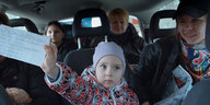 Aufnahme ins Innere eines Kleinbusses: Fünf Menschen sitzen auf den Rückbänken. In der Bildmitte sitzt ein Kind in einer rot-schwarz geblümten Winterjacke Jacke. Es trägt eine fliederfarbene Mütze und hält einen unleserlich beschrifteten Zettel in die Kam