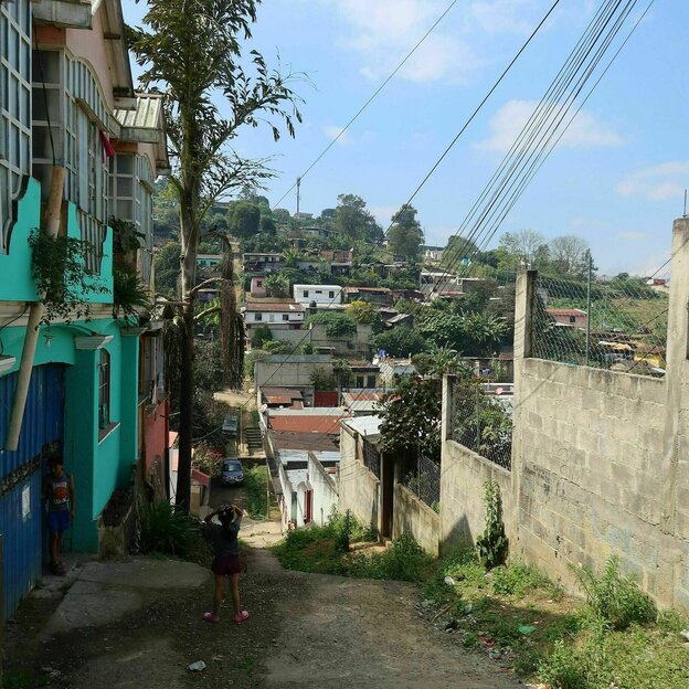 View of the district around the School of Hope, an unplastered wall stands on a steep, unpaved road, in front of brightly painted houses.