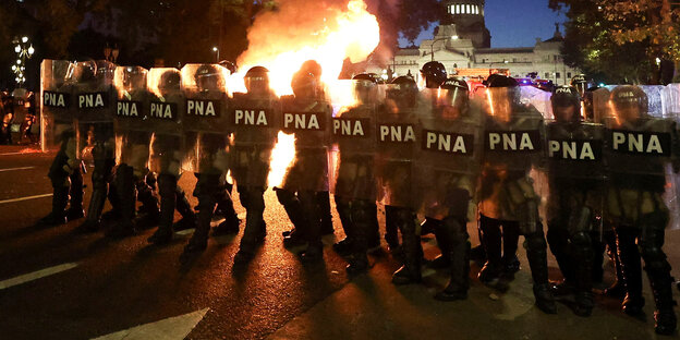 Uniformed people with protective shields and a fire burning behind them.