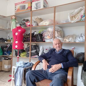 An elderly man sits in an armchair in a tailor's studio
