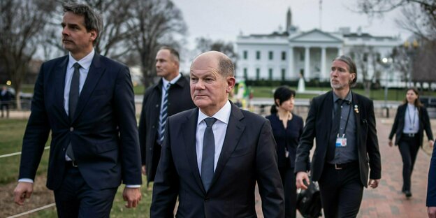 Chancellor Olaf Scholz (SPD) approaches the viewer with a group of employees and a serious face;  In the background you can see the White House.