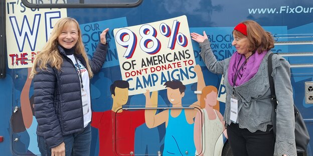 Two women stand in front of a blue campaign bus and point to promotional materials for Nikki Haley.