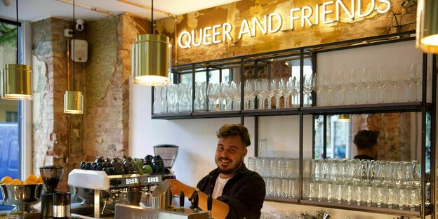 Danjel Zarte stands behind the counter and drinks a beer;  Above it, in big, bright letters it says: queer and friends.