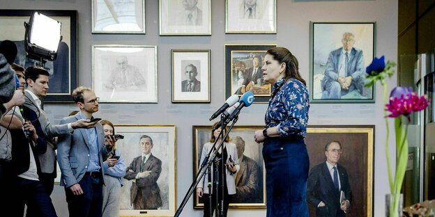 The outgoing Secretary of State for Culture and Media, Fleur Graper, speaks to the press.  On the walls there are numerous paintings depicting men.  She confronts journalists, mostly men.