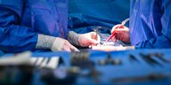 Zwei Ärzte führen eine Operation an einem Patienten in einem Krankenhaus durch. Der Patient sit völlig mit einem blauen Tuch abgedeckt, zu sehen sind nur behandschuhte Hände und Operationsbesteck