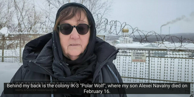 Alexei Navalny's mother Lyudmila Navalnaya stands in front of Charp prison, wearing sunglasses and a scarf around her head: barbed wire on a fence, there is snow
