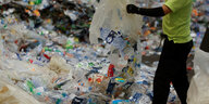 Ein arbeitender Mensch mit Schutzhandschuhen steht in einem Haufen leerer Plastikflaschen und kippt weitere Flaschen aus Plastiksäcken aus.