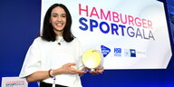 Reem Khamis bei der Ehrung zu Hamburgs Sportlerin des Jahres. Sie trägt offene, schulterlange schwarze Haare, ein weißes Shirt mit modisch-weiten Ärmeln und in der linken Hand den Preispokal: Eine Art runde Scheibe, durch die ein stilisiertes Hamburger Ra