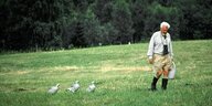 Konrad Lorenz als alter Mann. Er geht in Gummistiefeln über eine Wiese, gefolgt von drei Gänsen. Er trägt einen Eimer. Im Hintergrund Wald.