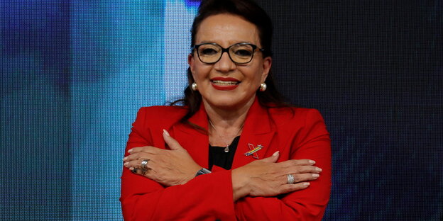 President Xiomara Castro.  She holds her upper arms with her hands, so that her arms are crossed.  She is wearing a red jacket.