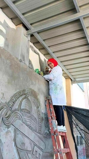 An image of happy days: Menna Hamouda, standing on a ladder, dressed in a painter's smock and a red headscarf, smiles at the camera.