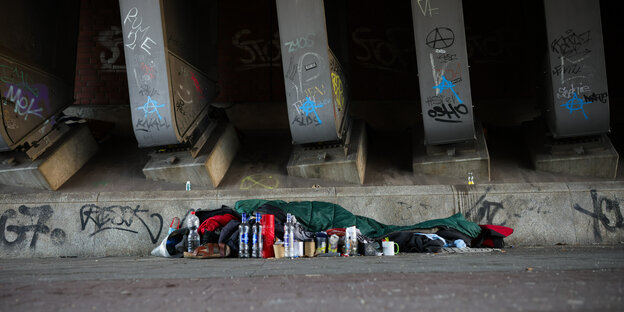 Ein Mensch liegt in einem grünen Schlafsack auf grauem Steinboden neben Brückenpfeilern.