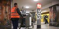 Eine Reinigungskraft, die eine Warnweste trägt, geht mit einem Reinigungswagen durch eine Beliner U-Bahn-Station.