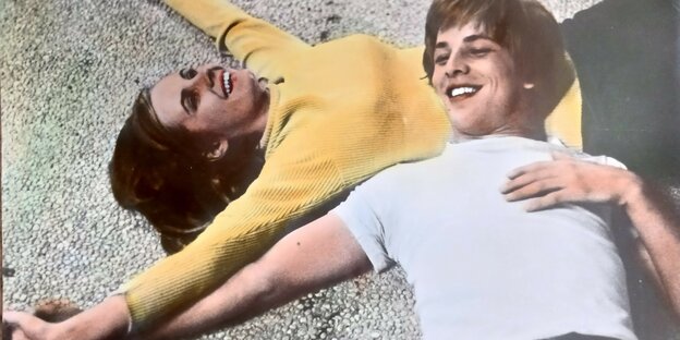 Eine junge Frau in gelbem Pulli und ein junger Mann in hellem T-Shirt liegen auf dem Boden und lachen. Sie halten sich an den Händen, der Mann ruht mit dem Kopf auf dem Bauch der Frau.