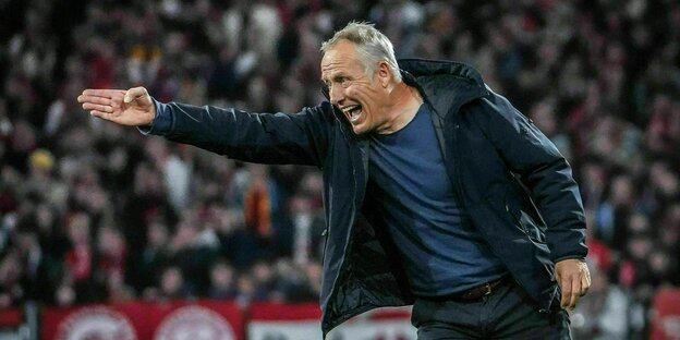 Coach Christian Streich points his arm to the left on the soccer field while shouting