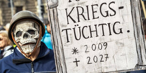 Demoschild in Form eines Grabsteines, auf dem steht "Ich war kriegstüchtig". Daneben ein Demonstrant mit Totenkopfmaske. Anti-Siko-Demo am Stachus, München, 17. Februar a2024