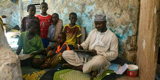 Teacher Abas M'Ballo is sitting on the floor in bright traditional clothing and holding a blackboard, and six children sit next to him and listen to him.