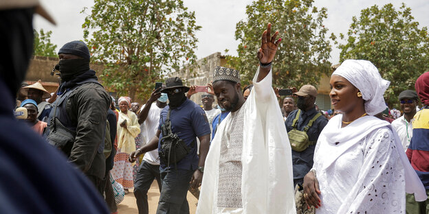 Bassirou Diomaye Faye im weißen Gewand umgeben von vermummten Personenschützern, winkt