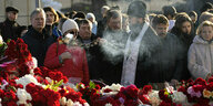 Ein orthodoxer Priester steht vor einem Blumenmeer, umringt von einer Menschenmenge und schwenkt einen Weihrauchkessel