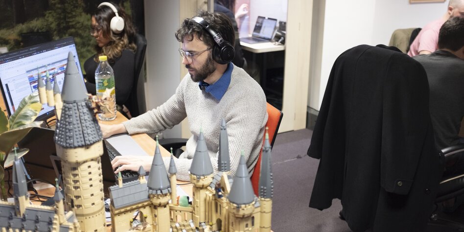 Ein junger Mann sitzt mit Kopfhörern vor einem Monitor, auf seinem Schreibtisch steht eine Lego-Zauberschule