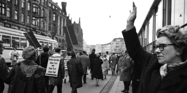 Black and white photo, a woman stands at the edge of a demonstration with her hand raised.  Participants were photographed from behind;  One of them carries a sign on his back: There is no bomb without testing, there is no testing without death.