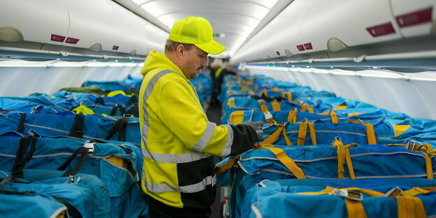 A postal worker stands in the hold of an airplane surrounded by blue mail bags.