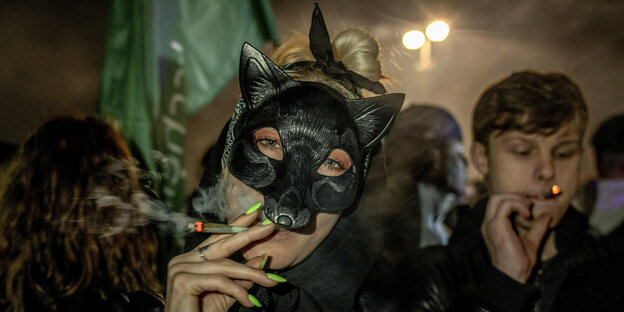 Eine Frau mit einer Wolfsmaske und langen grünen Fingernägeln zieht an einem Joint.